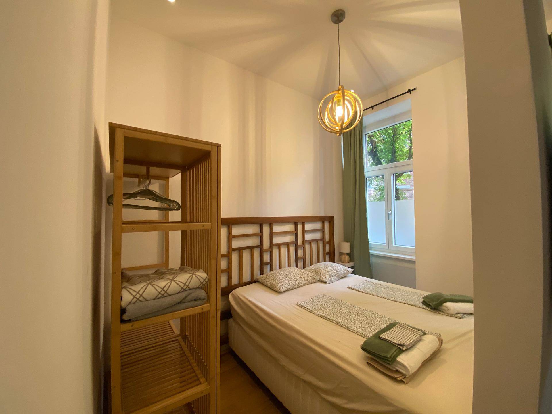 Schlafzimmer mit Holzbettrahmen, offenem Regal und Fenster mit grünen Vorhängen.