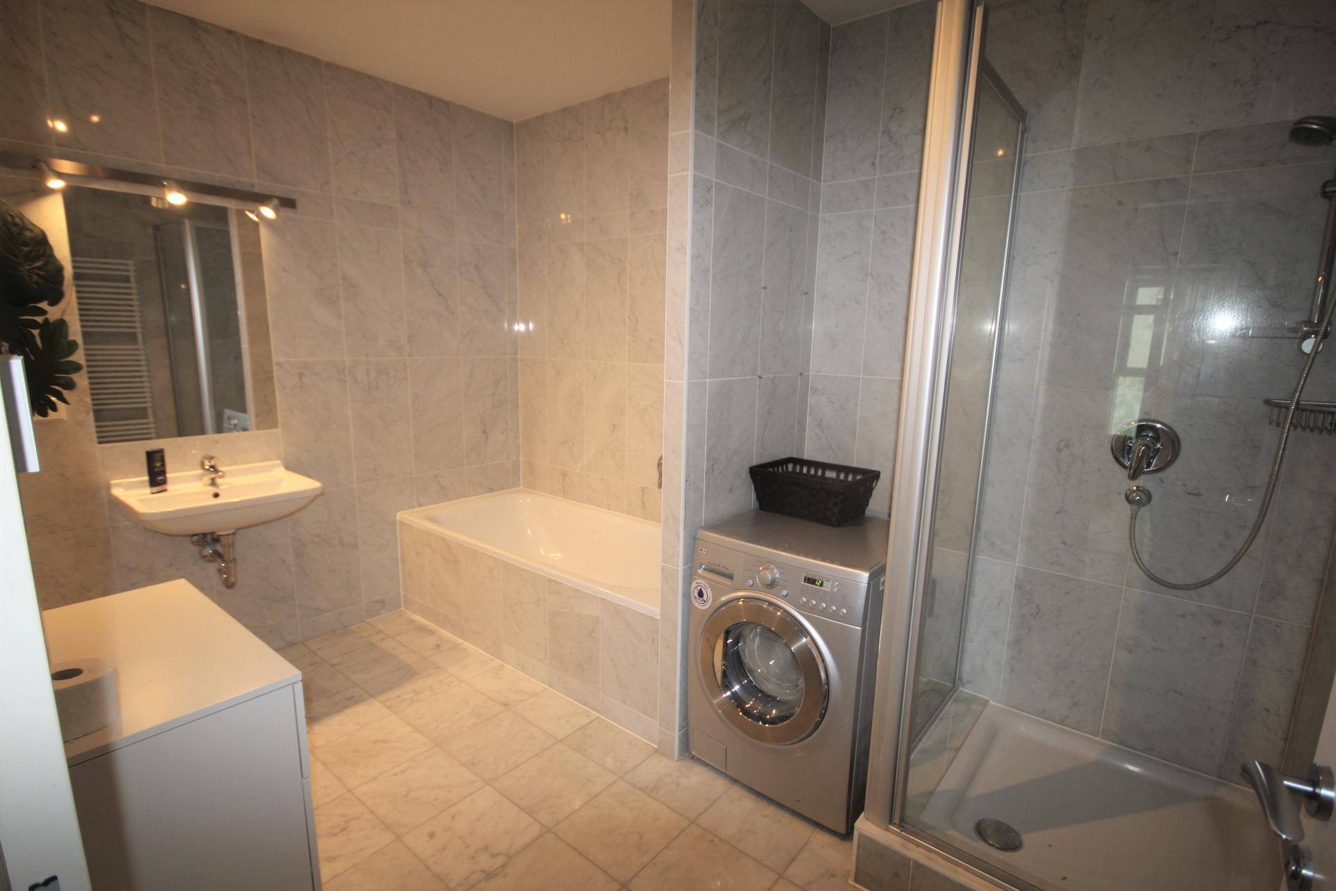 Modernes Badezimmer mit hellgrauen Fliesen, Badewanne, Dusche und Waschmaschine.