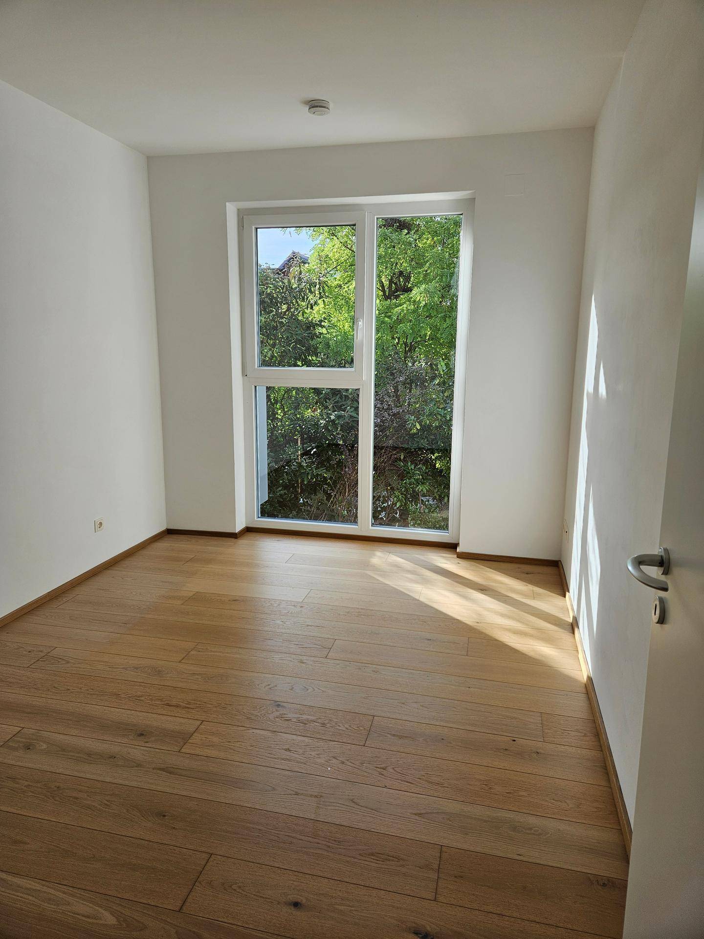 Helles Zimmer mit Holzboden und großem Fenster, das einen Blick ins Grüne bietet.