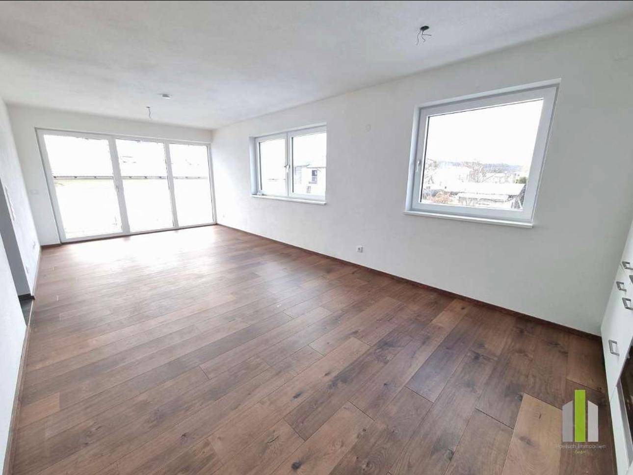 Geräumiger Wohnbereich mit dunklem Holzboden und großen Fenstern, die viel Tageslicht hereinlassen.