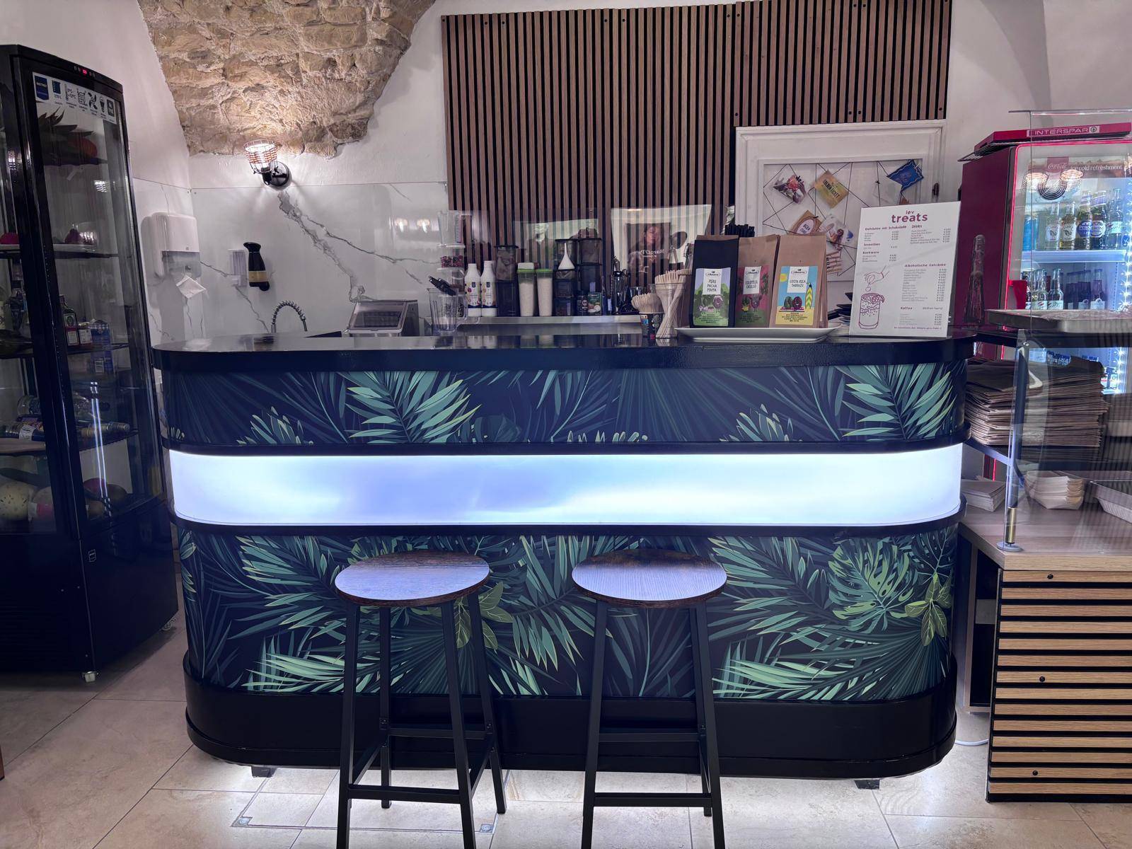 Moderne Theke mit tropischem Blattmuster, integrierter Beleuchtung und zwei Barhockern im Café.