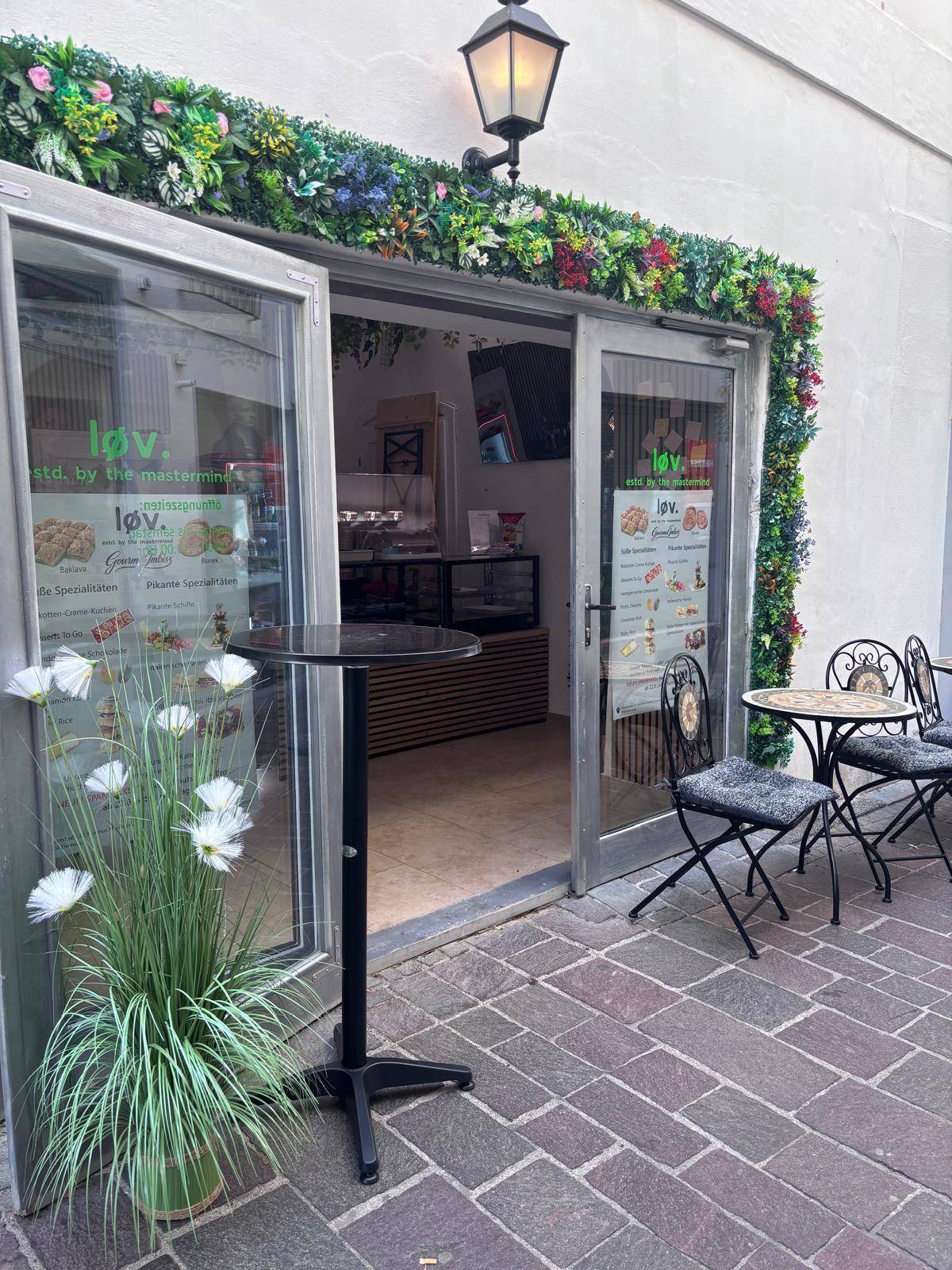 Café-Eingang mit geöffneten Glastüren, Außentischen und grüner Girlande über dem Rahmen.