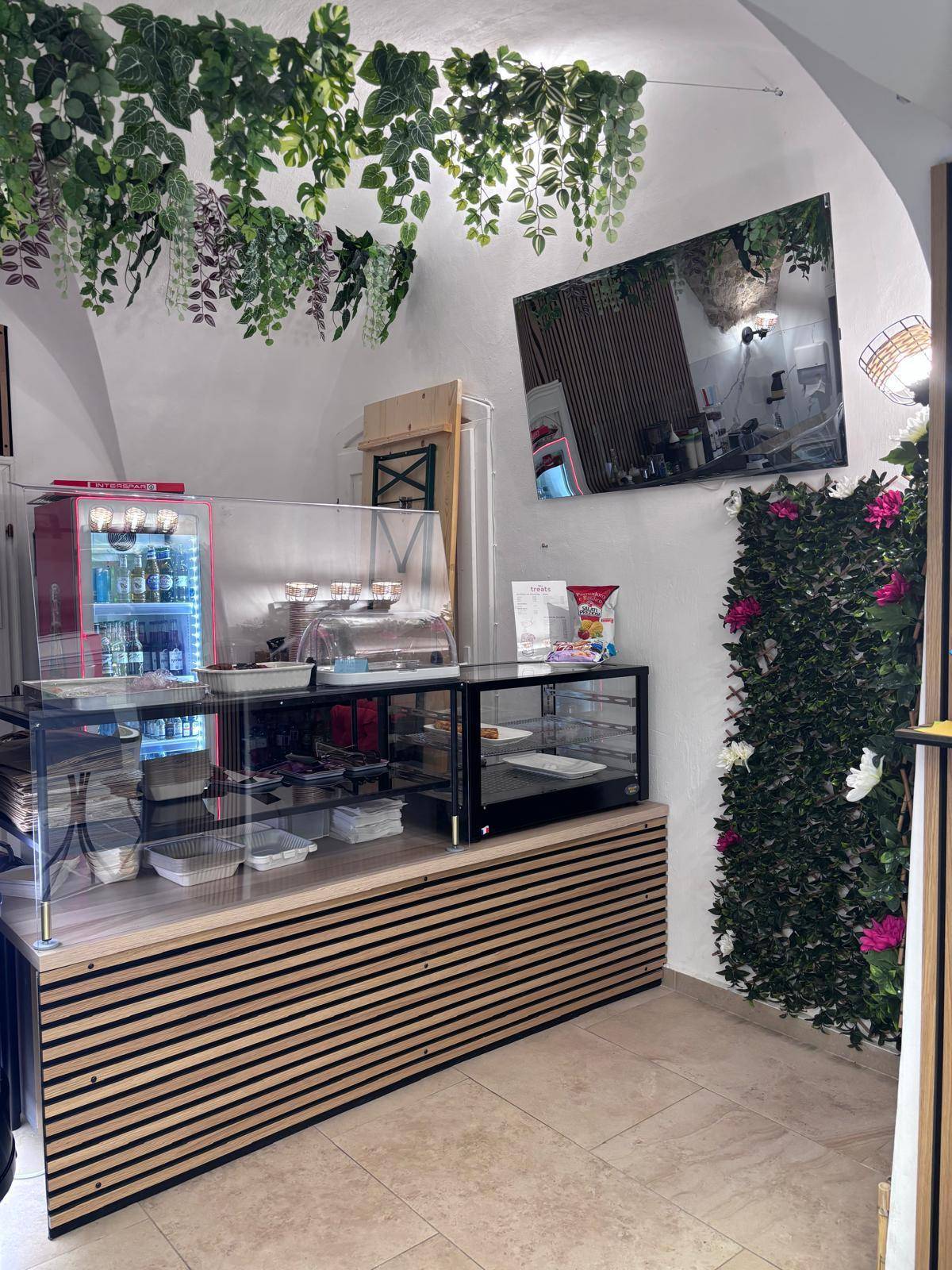 Innenansicht eines Cafés mit Holzlamellen-Theke, Vitrinen und hängenden Kunstpflanzen an der Decke.
