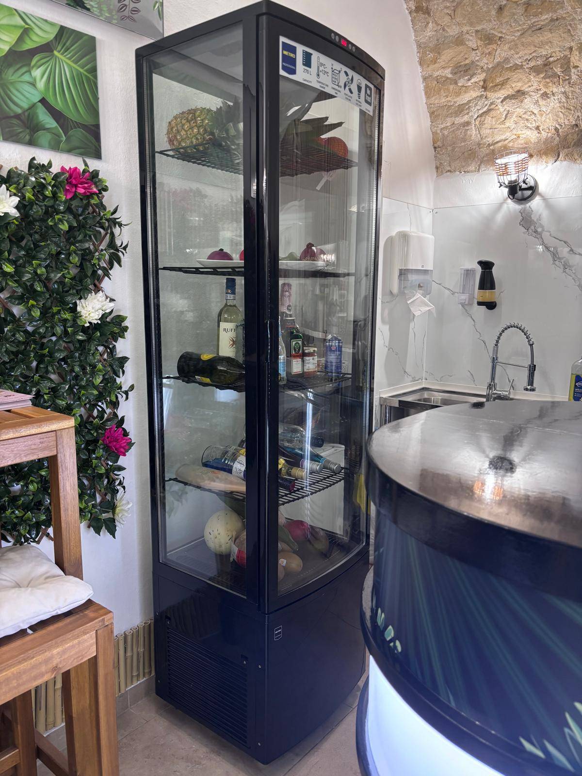 Hoher Kühlschrank mit Glasfront, gefüllt mit Getränken und Produkten, neben der Theke.
