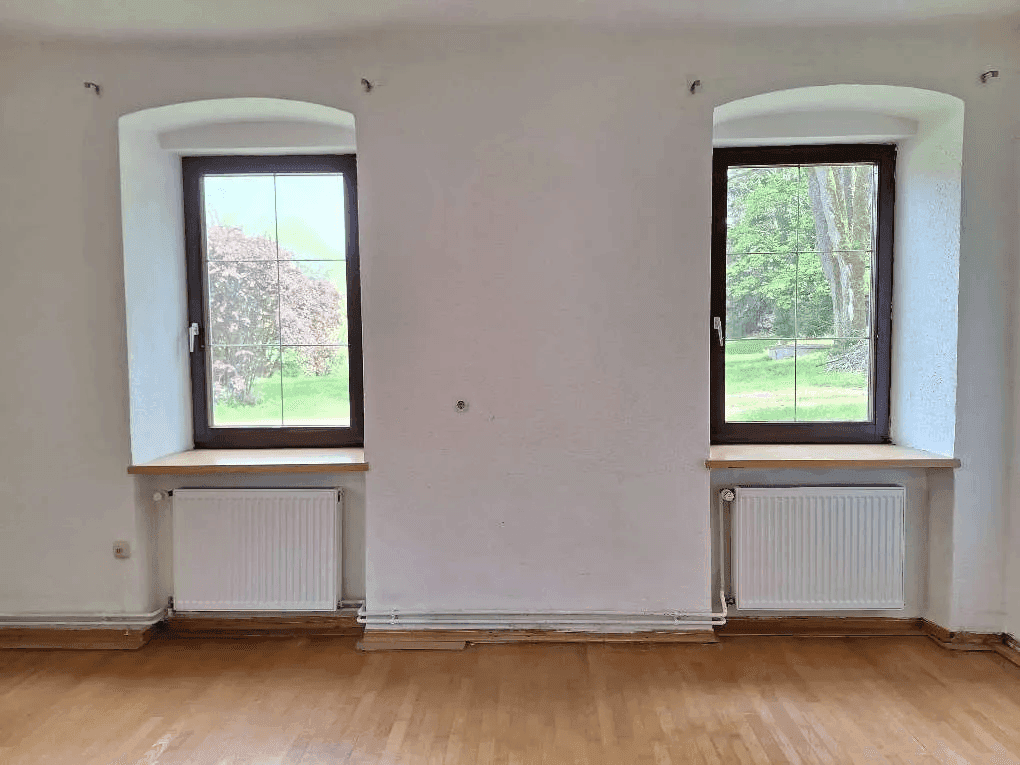 Helles Zimmer mit zwei Fenstern, Holzboden und weiß gestrichenen Wänden.