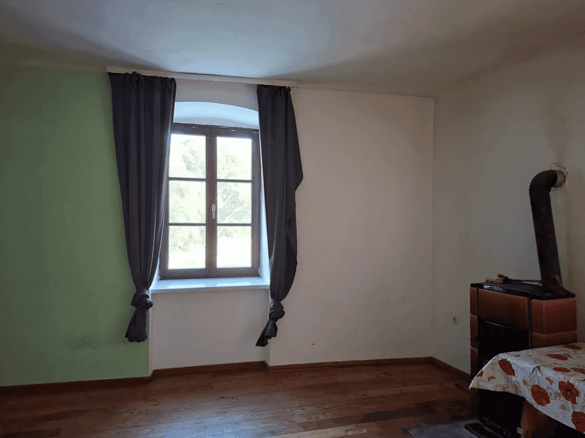 Helles Zimmer mit Holzboden, Kaminofen und einem Fenster mit Blick ins Grüne.