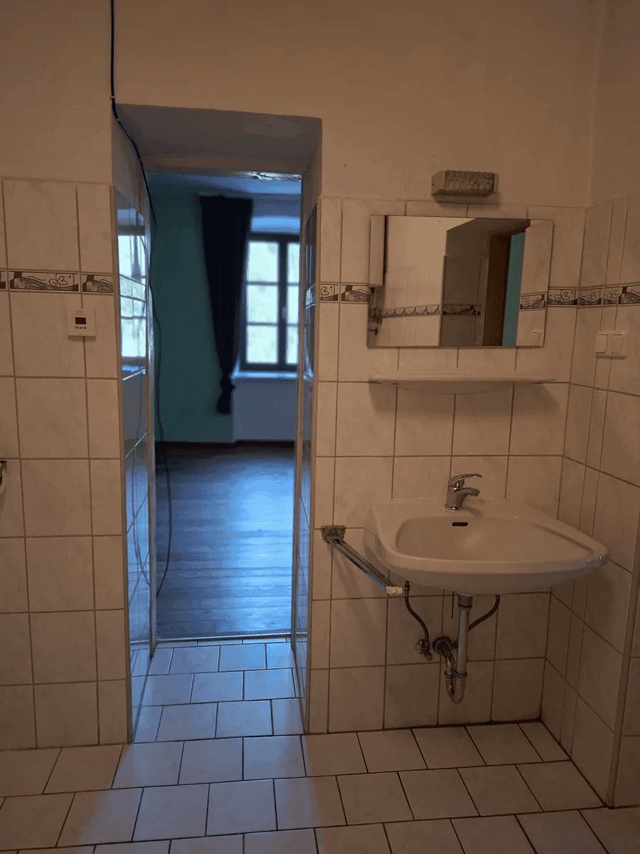 Einfaches Badezimmer mit Waschbecken und Fliesen, das zu einem weiteren Raum führt.