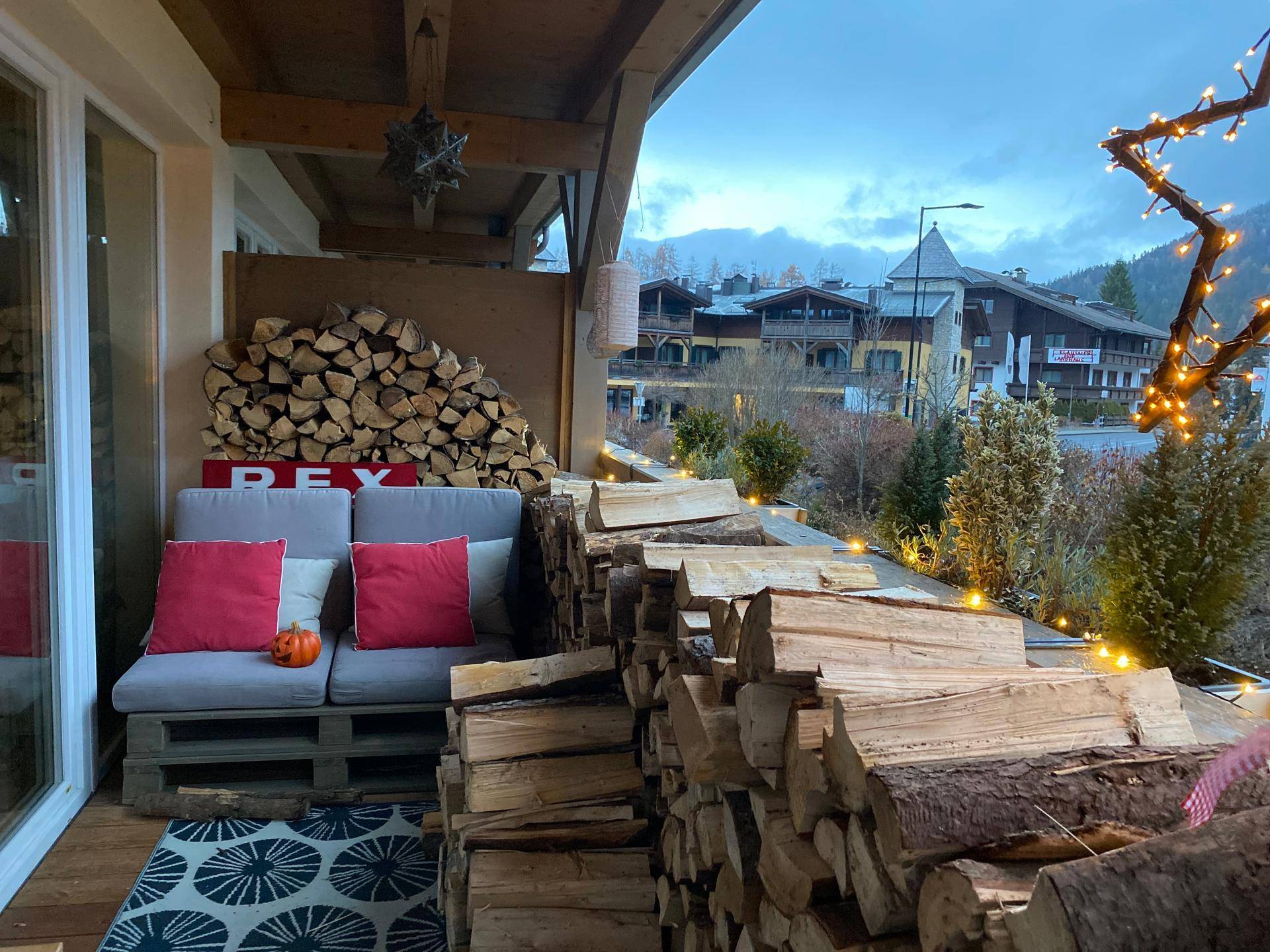 Überdachter Balkon mit Holzstapeln, gemütlicher Sitzecke und Blick auf die beleuchtete Umgebung.