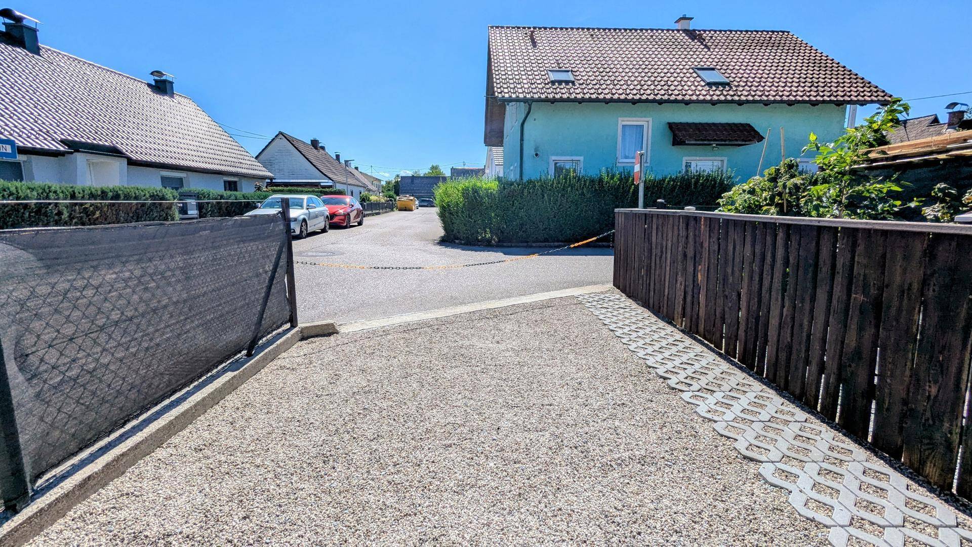 Blick von einem Kiesparkplatz auf die Wohnstraße mit einem türkisfarbenen Haus und weiteren Gebäuden im Hintergrund.
