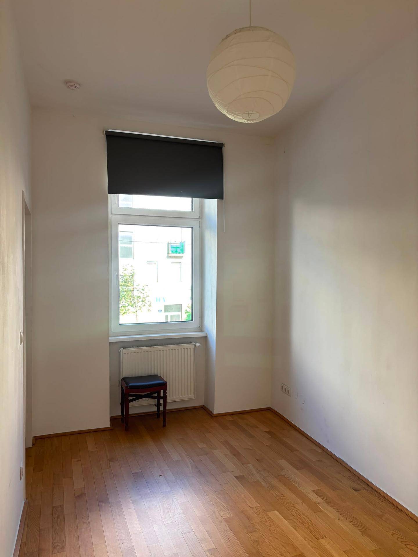 Leerer Raum mit Holzfußboden, Fenster mit Rollo und Heizkörper unter dem Fenster.