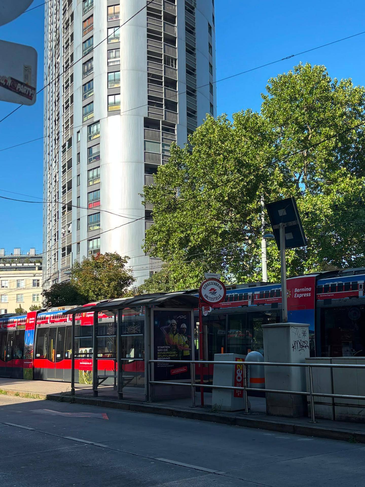 Städtische Szene mit Straßenbahnhaltestelle, hohem Gebäude und Bäumen an einem sonnigen Tag.