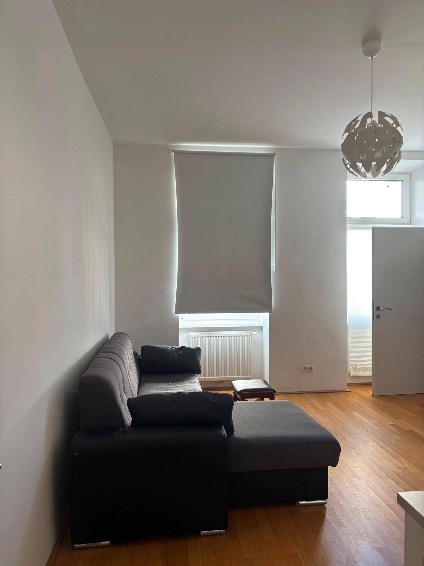 Wohnzimmer mit dunklem Sofa, Holzfußboden und einem Fenster mit Rollo.