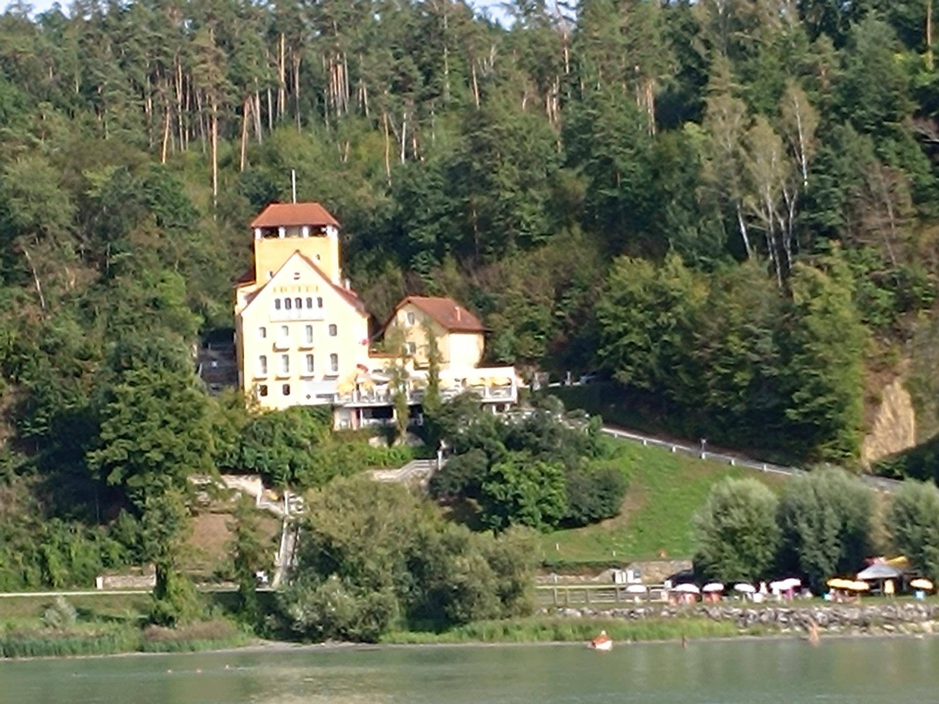 Panoramablick auf das historische Gebäude mit Turm auf dem Hügel, umgeben von Wald und dem See.