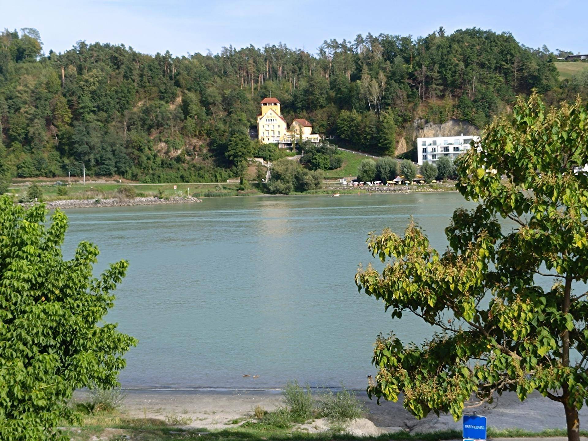 Weitläufiger Blick über den See auf die bewaldeten Hügel und ein markantes Gebäude am Ufer.
