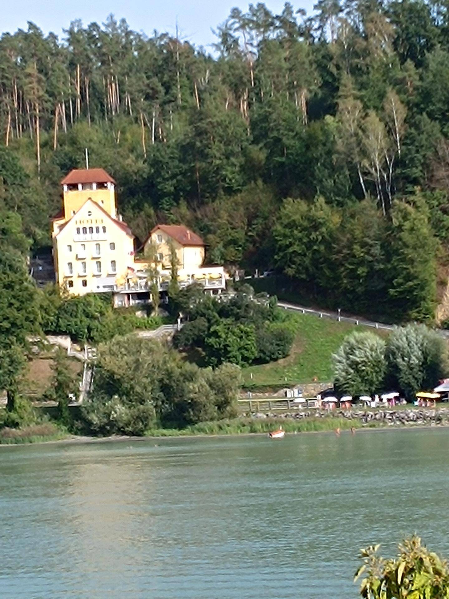 Nahaufnahme des gelben Gebäudes mit Turm auf dem bewaldeten Hügel, mit dem See im Vordergrund.