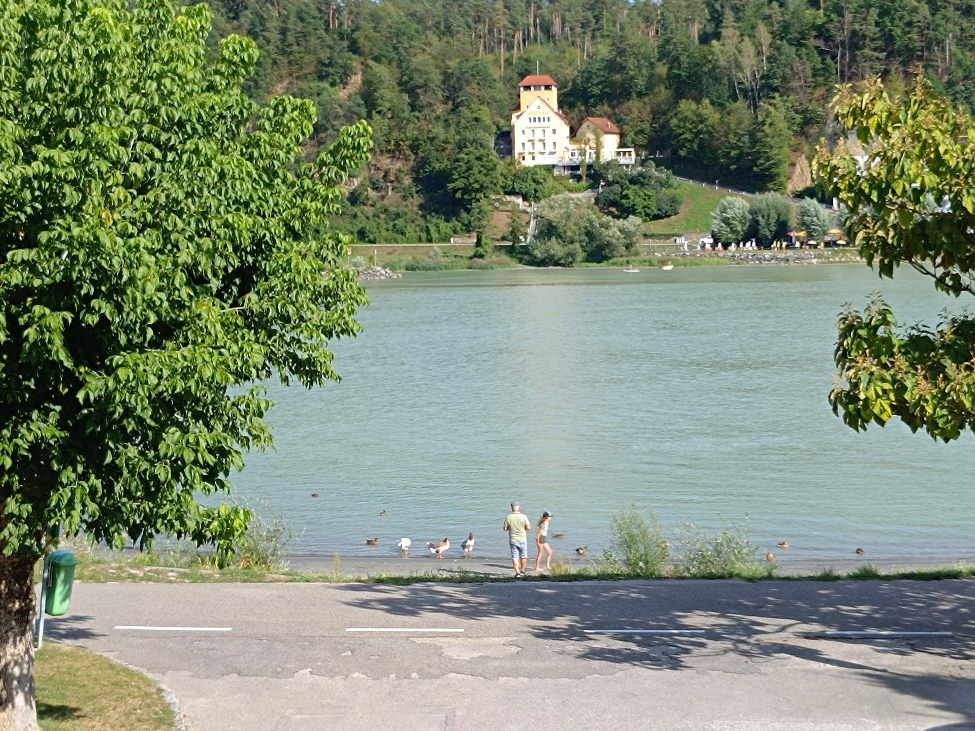Idyllischer Blick auf den See mit Menschen am Ufer und einem markanten Gebäude auf dem bewaldeten Hügel.
