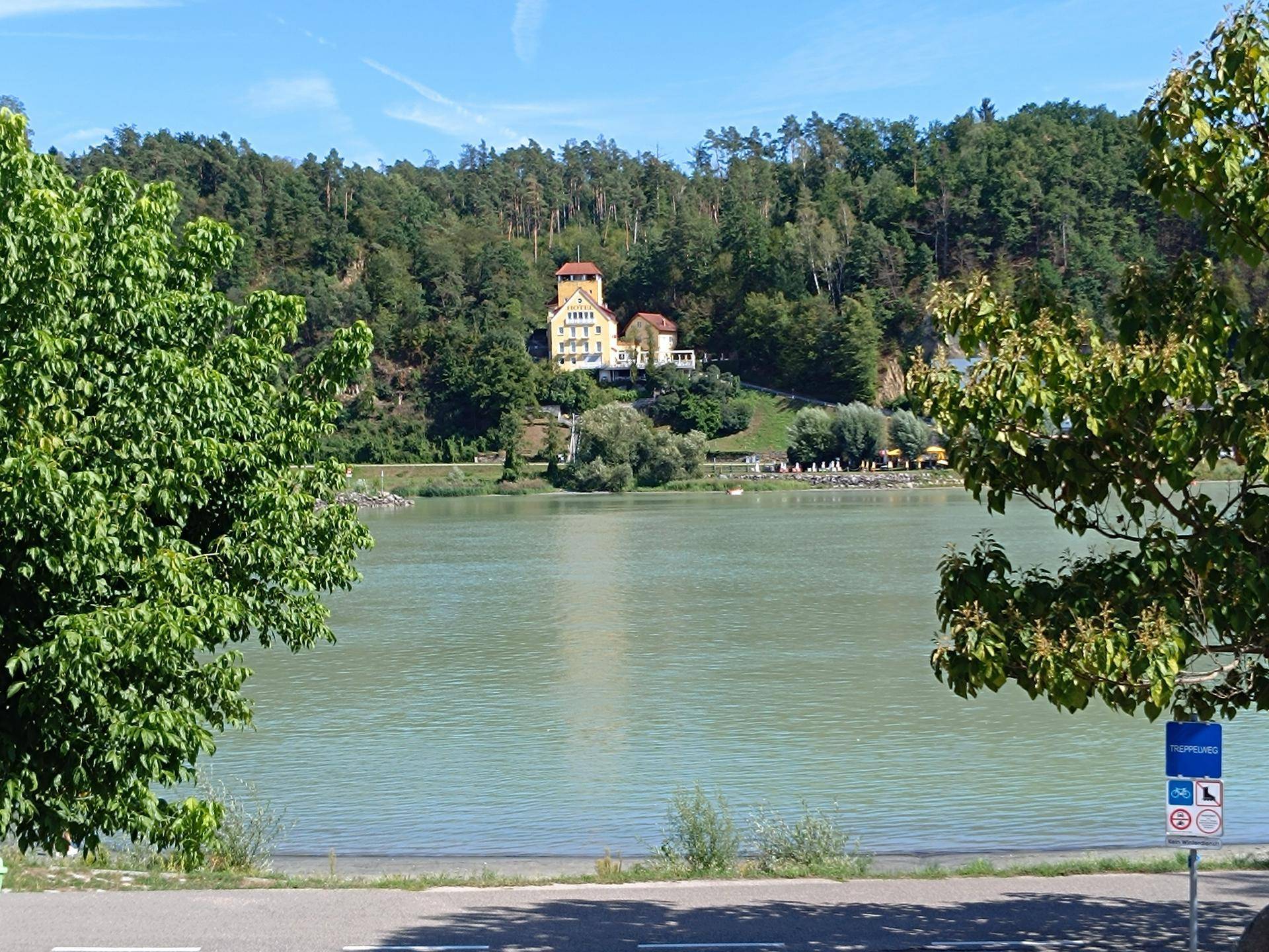 Panoramablick auf einen ruhigen See mit einem historischen Gebäude auf einem bewaldeten Hügel im Hintergrund.