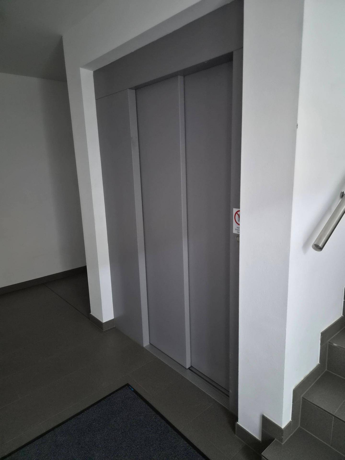 Heller Eingangsbereich mit modernem Aufzug und Treppenhaus in einem Mehrfamilienhaus.