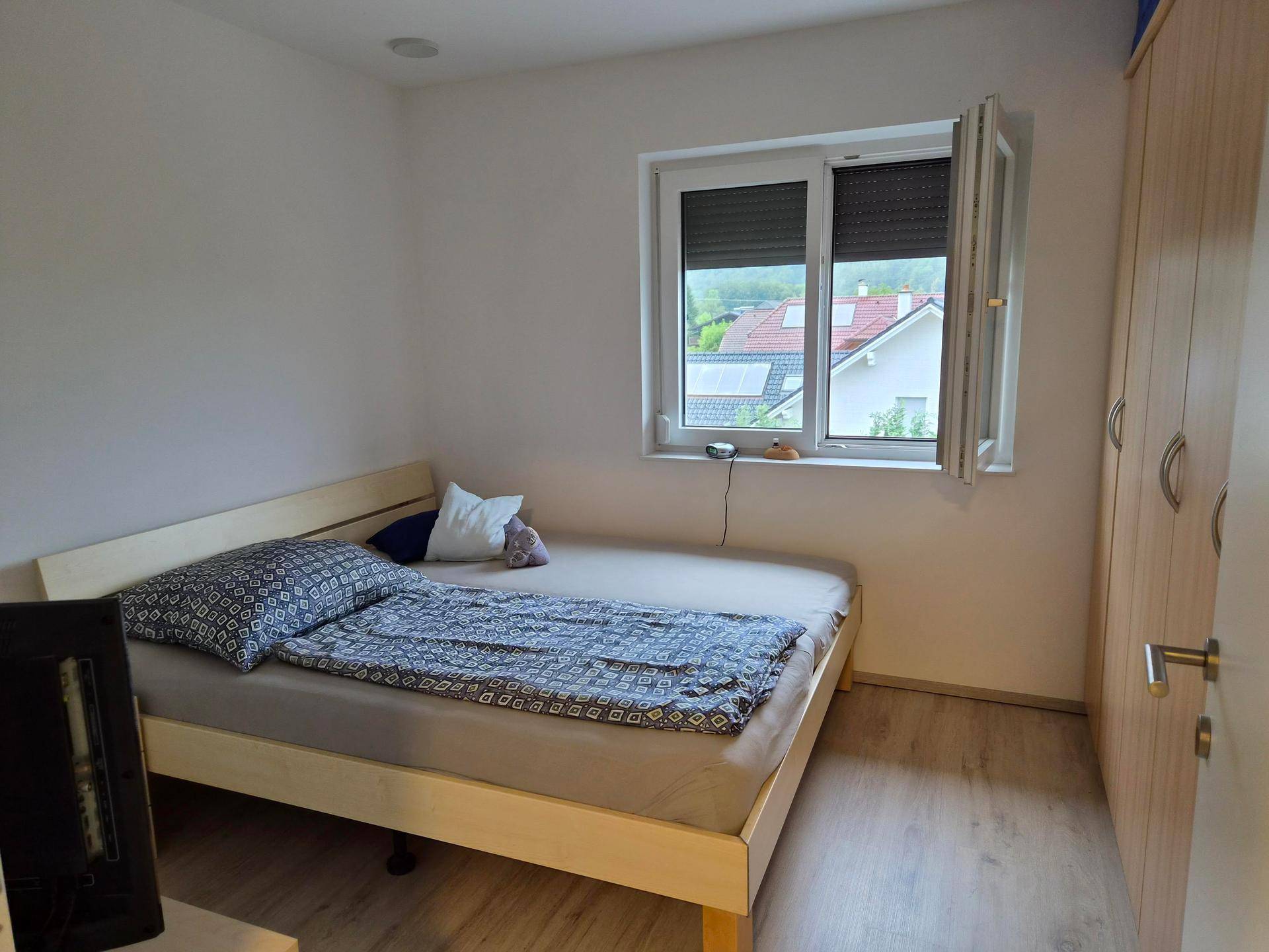 Helles Schlafzimmer mit einfachem Bett, Laminatboden und Fenster mit Rollladen.