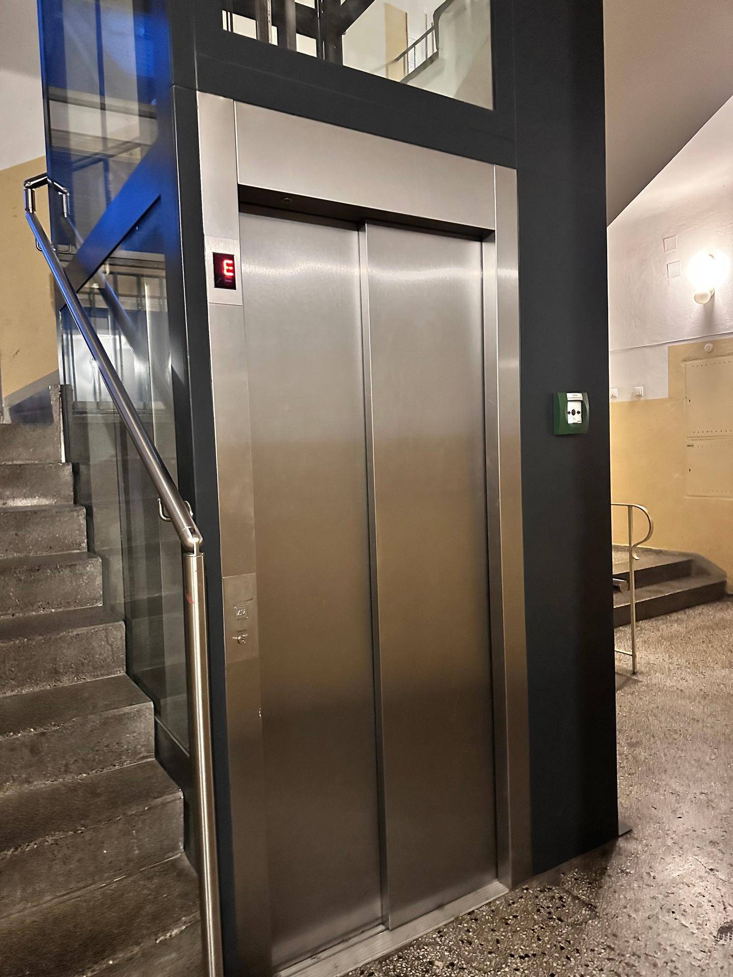 Moderner Aufzug im Treppenhaus eines Mehrfamilienhauses, mit Edelstahlfront und Glas.