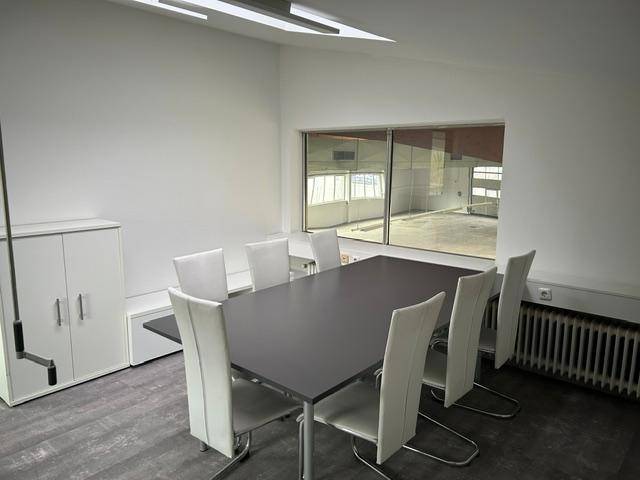 Besprechungsraum mit großem Tisch, Stühlen und Blick in eine angrenzende Halle.