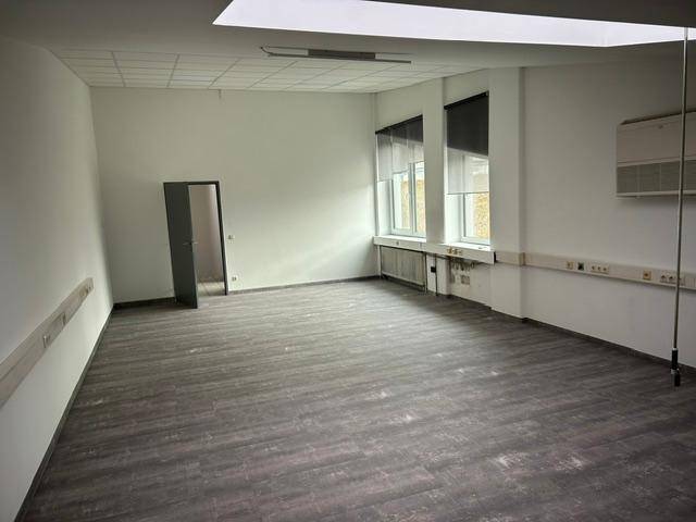 Leerer Büroraum mit modernem Laminatboden, weißen Wänden und Fenstern mit Jalousien.