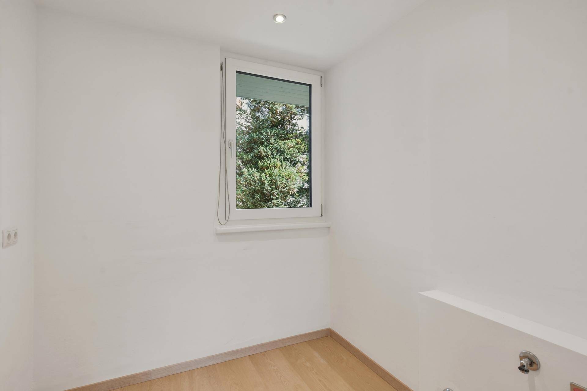 Helles Zimmer mit Holzboden und einem Fenster mit Blick ins Grüne.