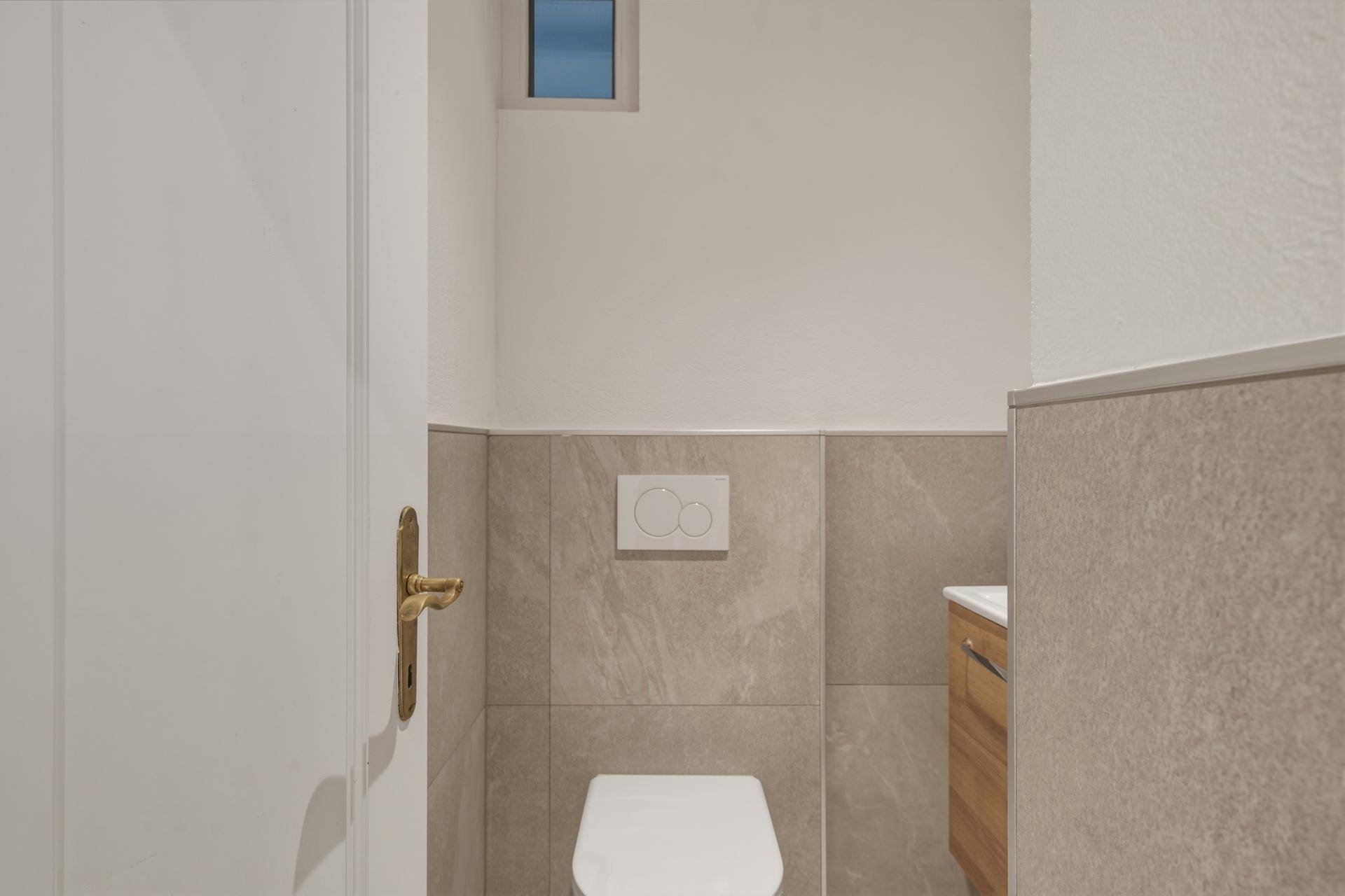 Separate Toilette mit wandhängendem WC, grauen Fliesen und kleinem Fenster.