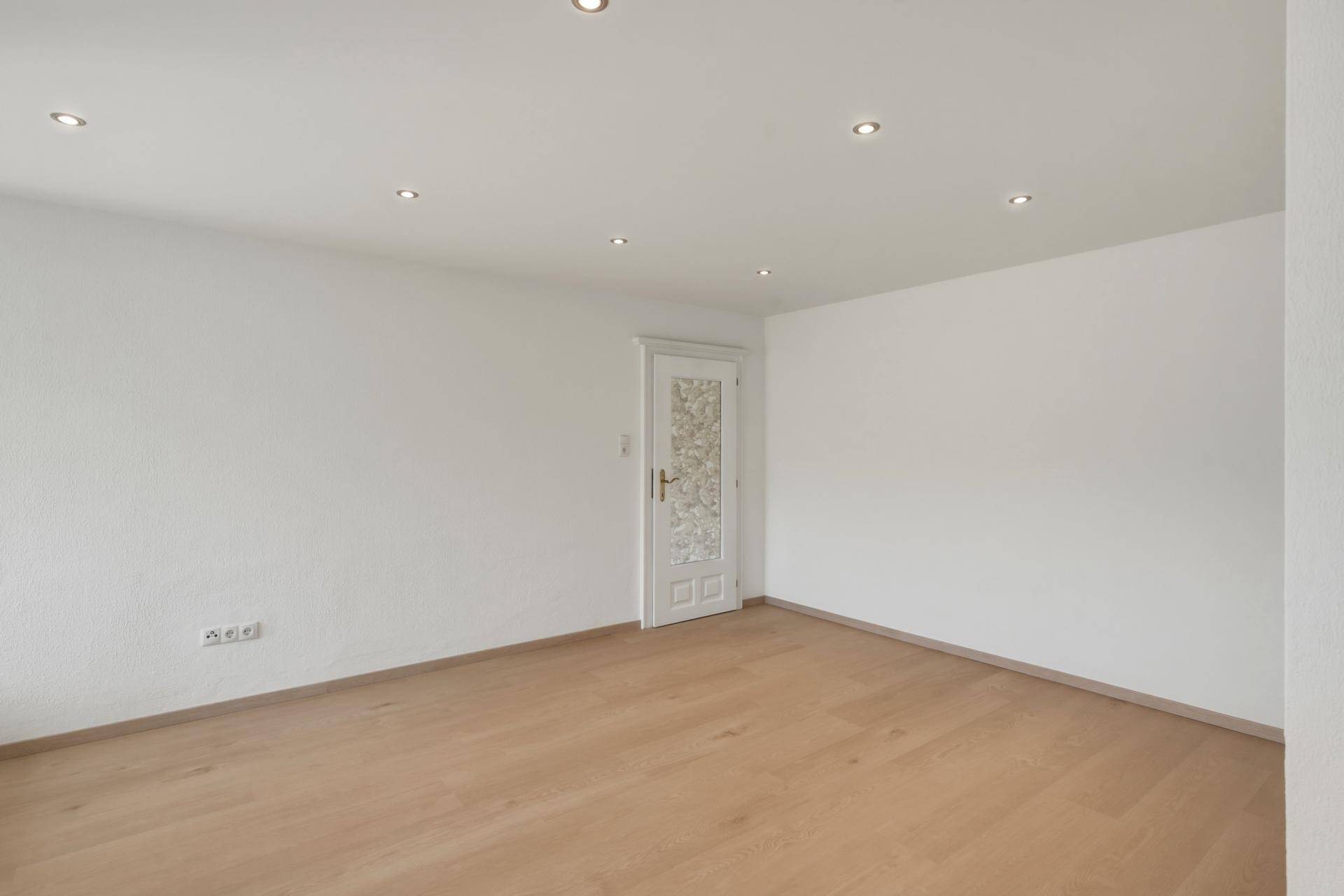Leeres Zimmer mit hellem Holzboden und einer Tür mit dekorativem Glaseinsatz.