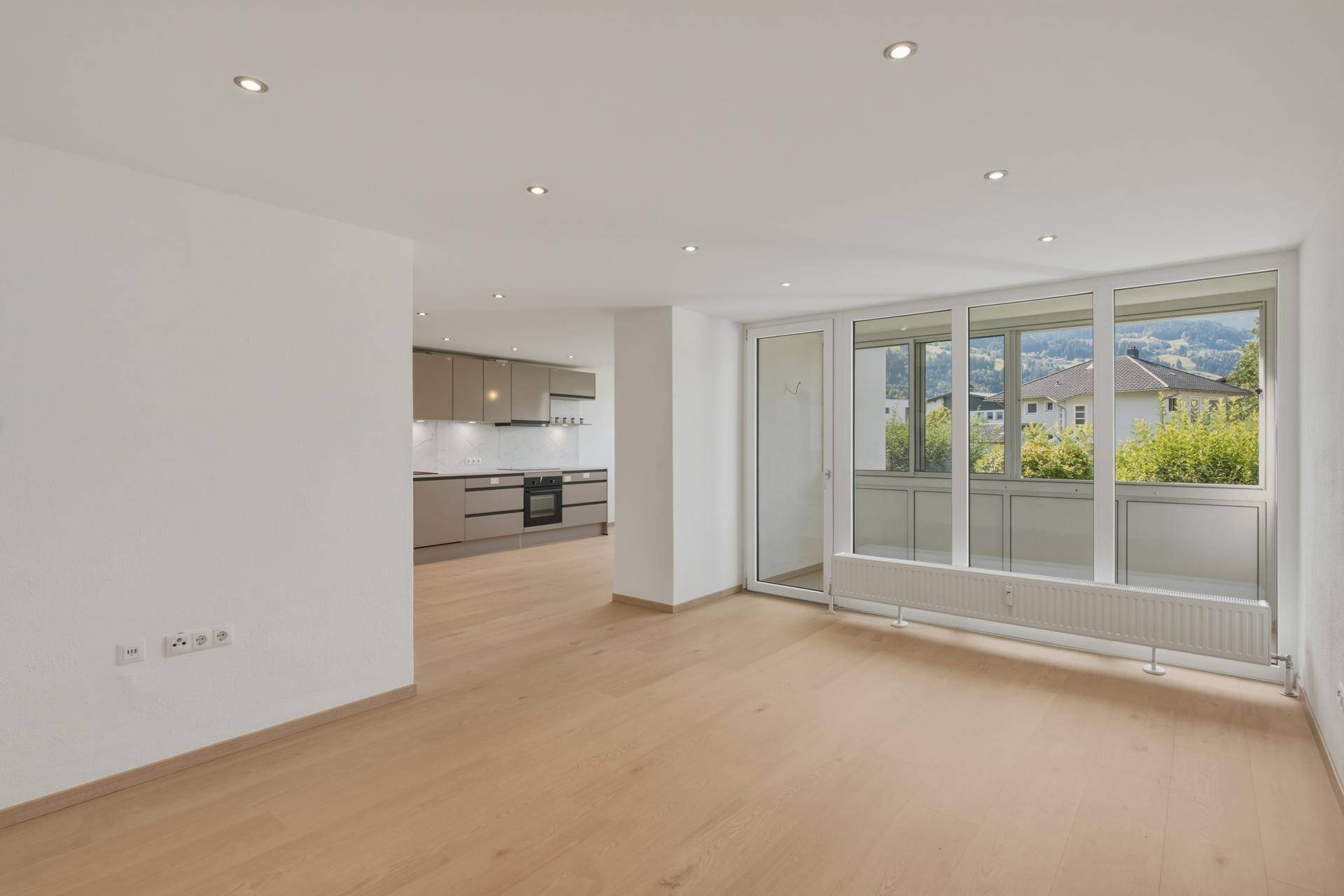 Offener Wohnbereich mit Holzboden, großen Fenstern und Zugang zu einem Balkon mit Bergblick.