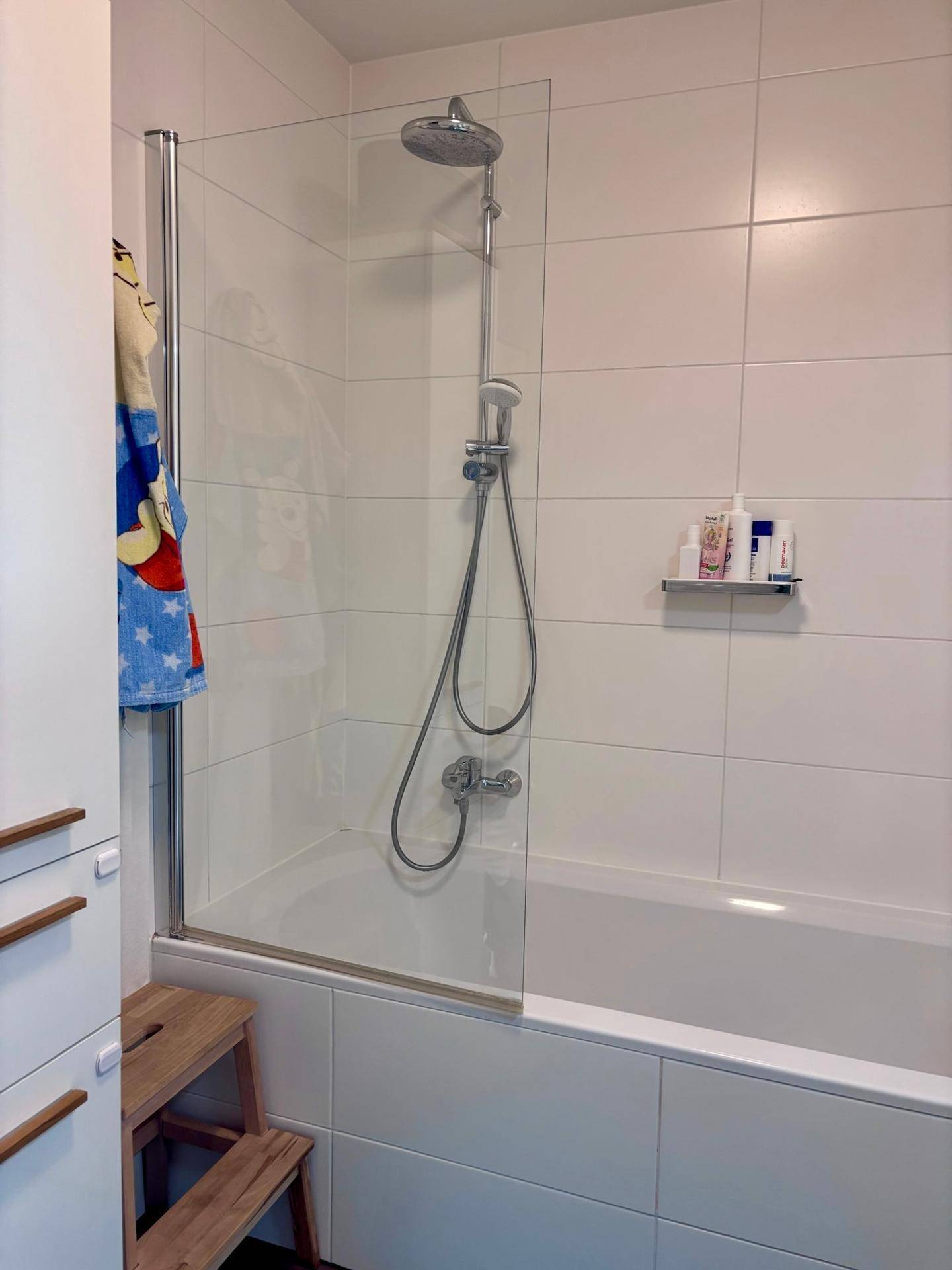 Badezimmer mit Badewanne und Duschvorrichtung, komplett gefliest in Weiß.