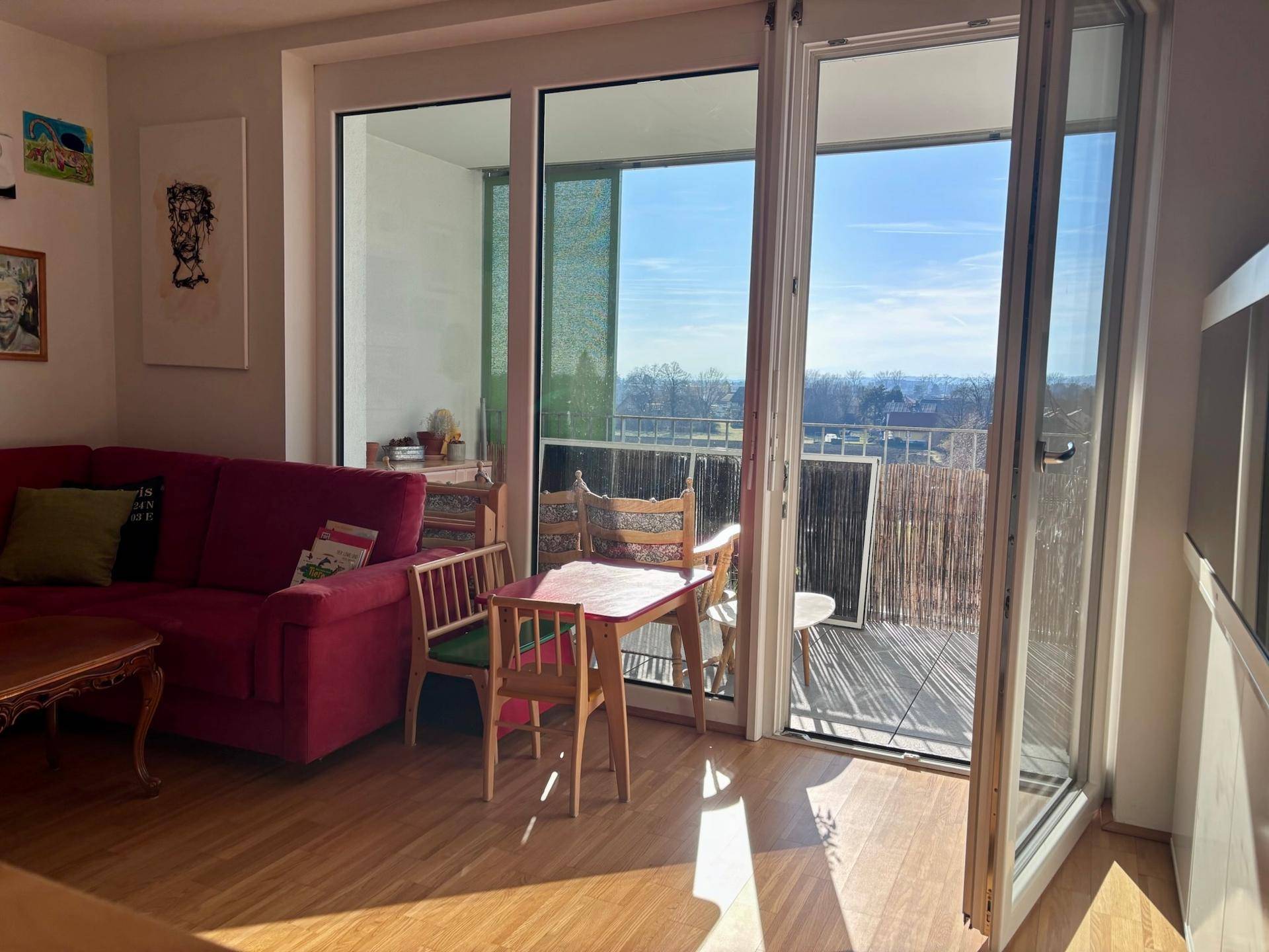 Helles Wohnzimmer mit rotem Sofa, Holzboden und Zugang zu einem sonnigen Balkon mit Weitblick.