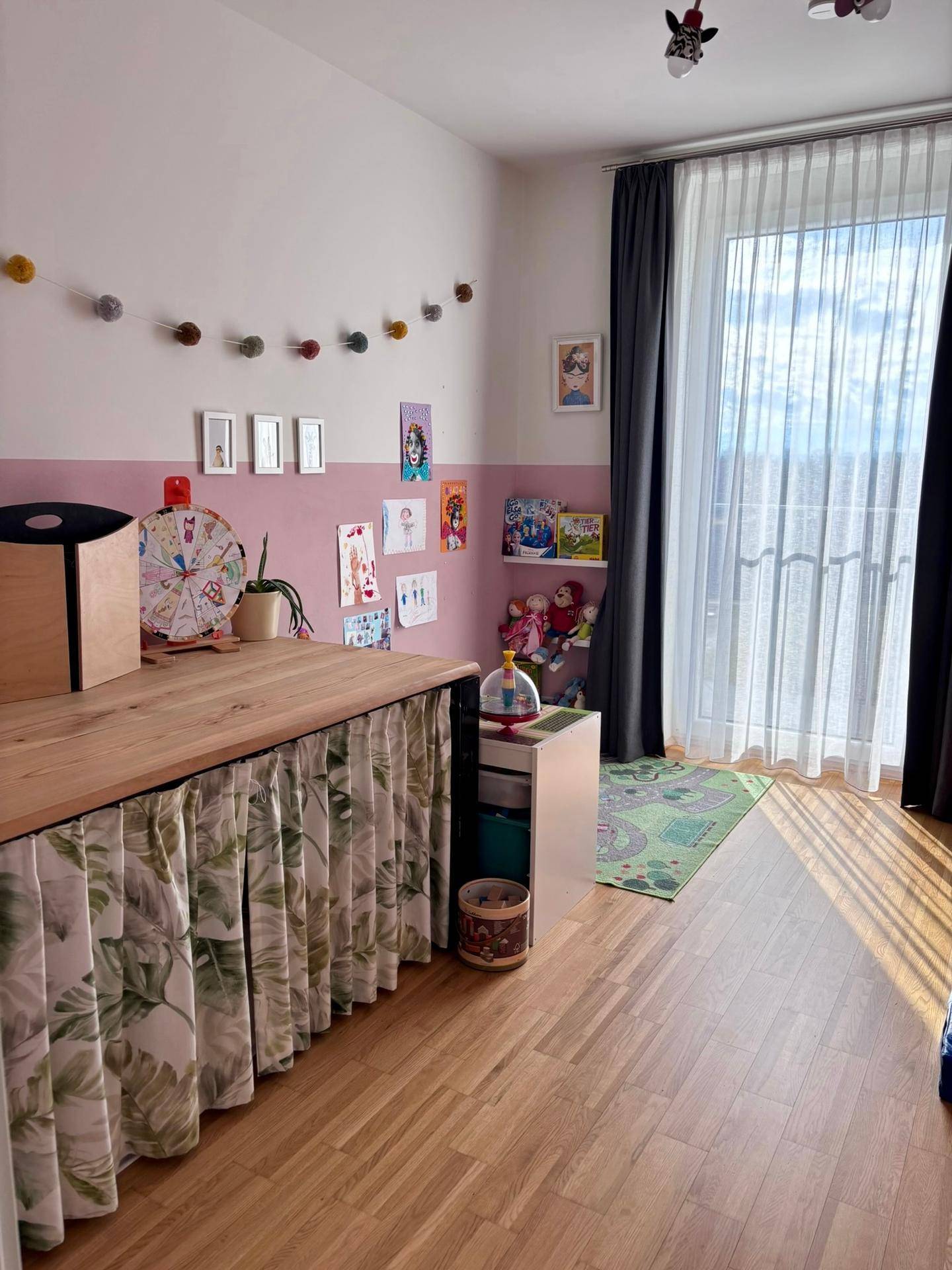 Kinderzimmer mit rosa Akzentwand, Holzmöbeln und einem Fenster mit Vorhängen.