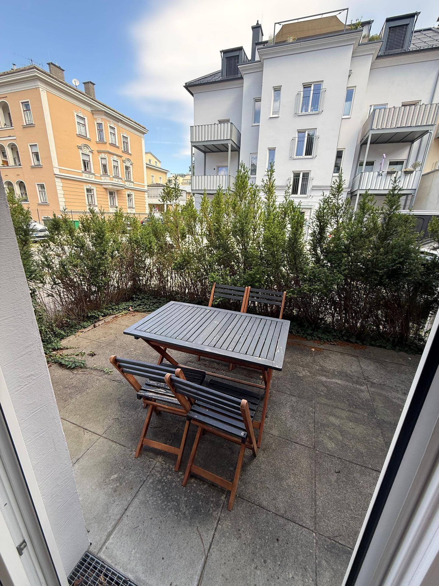 Gemütliche Terrasse mit Holztisch und Stühlen, umgeben von Hecken, ideal für Mahlzeiten im Freien.