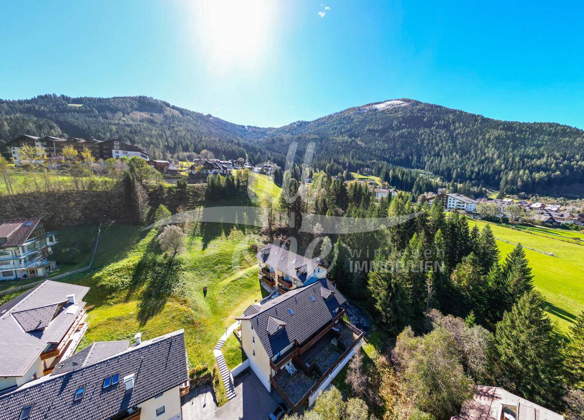 Luftaufnahme eines Anwesens in einer grünen Berglandschaft unter strahlend blauem Himmel.