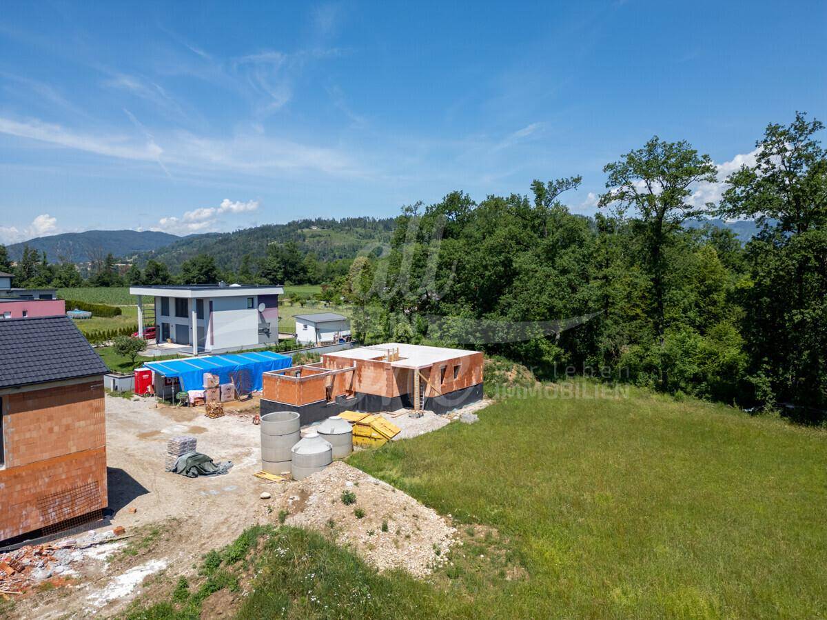 Blick auf ein im Bau befindliches Haus mit Rohbau und grüner Umgebung unter blauem Himmel.