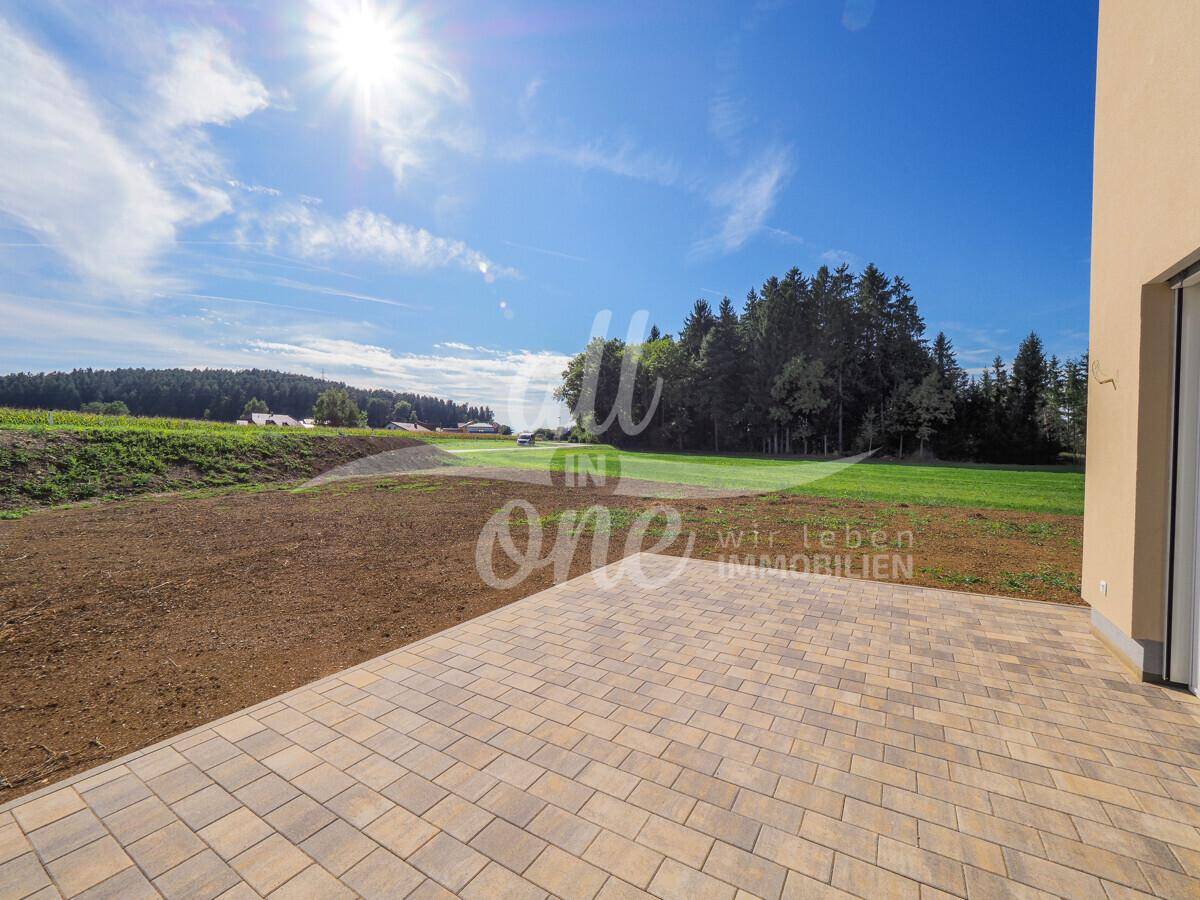 Gepflasterte Terrasse neben dem Haus mit weitem Blick auf die grüne Landschaft unter blauem Himmel.