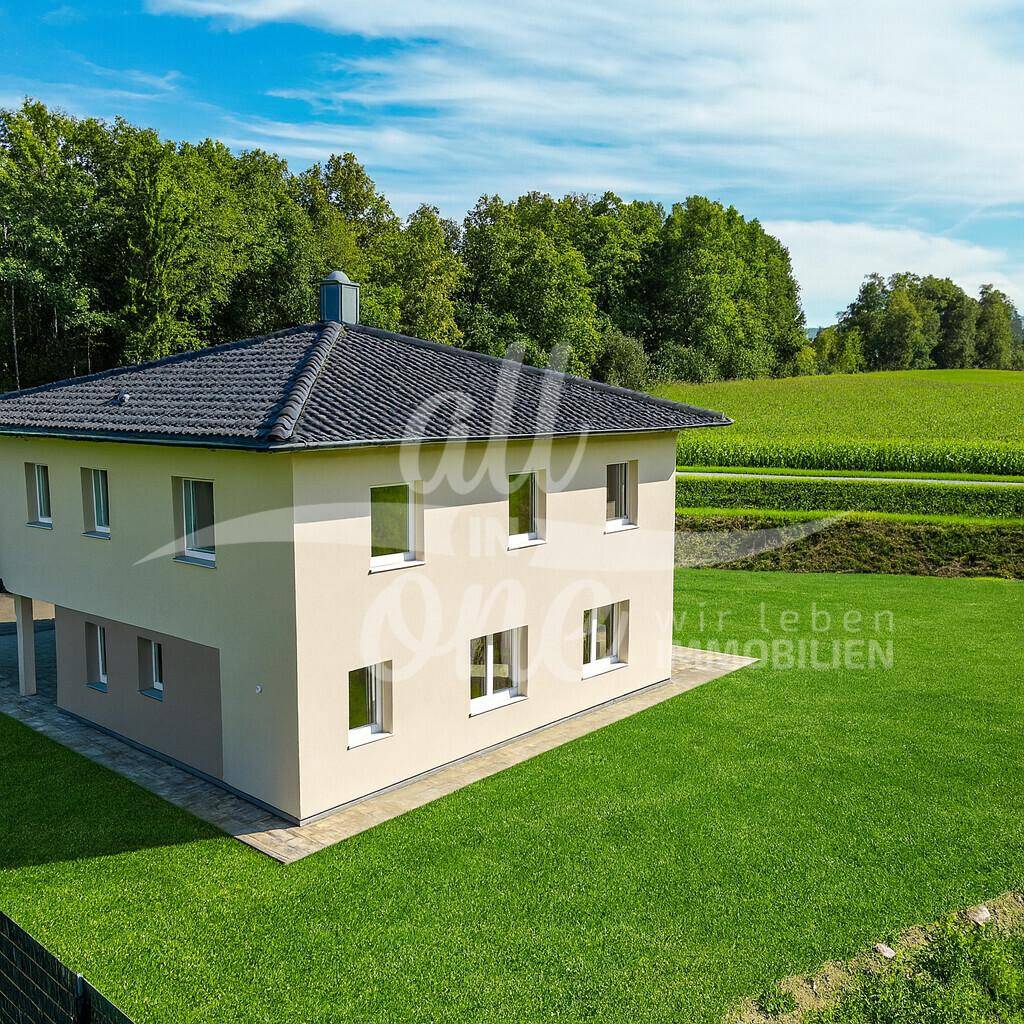 Modernes Haus umgeben von grünen Feldern und Wald, mit gepflegtem Rasen und blauem Himmel.