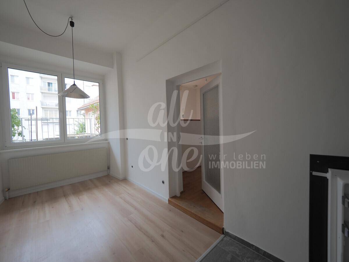 Heller Wohnbereich mit großem Fenster und hellem Laminatboden, angrenzend an die Küche.