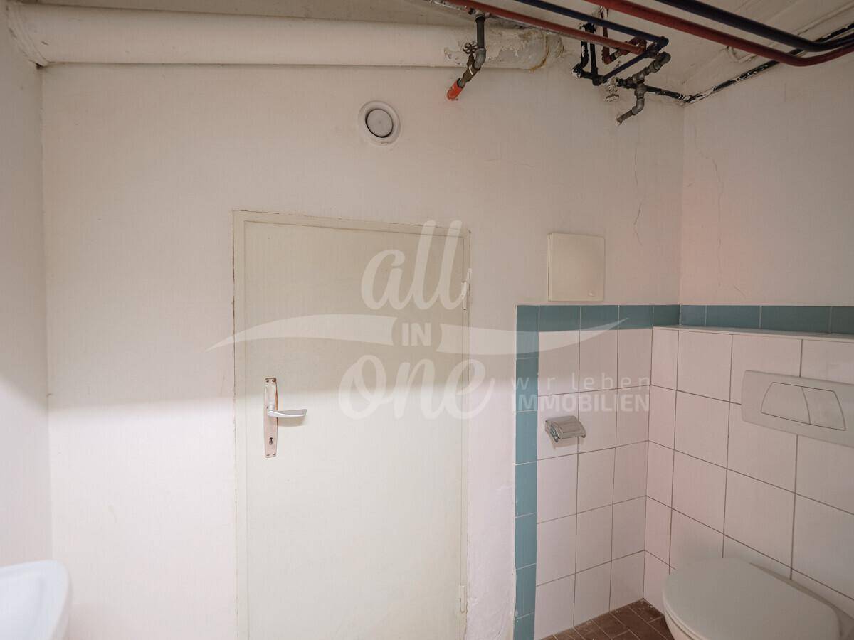 Einfaches WC mit Fliesen im Stil der 80er Jahre, sichtbaren Leitungen und einem einzelnen Waschbecken.