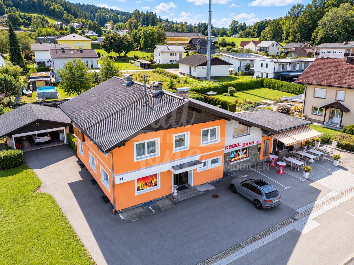 Luftaufnahme des Gebäudes mit umliegenden Häusern, Parkplätzen und grüner Hügellandschaft im Hintergrund.