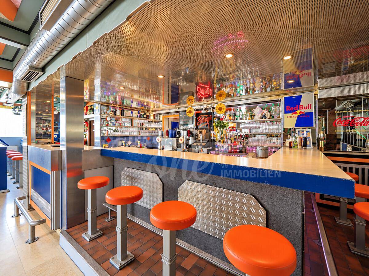 Detailansicht einer Bar mit orangefarbenen Hockern, glänzenden Oberflächen und einer gut bestückten Rückwand.