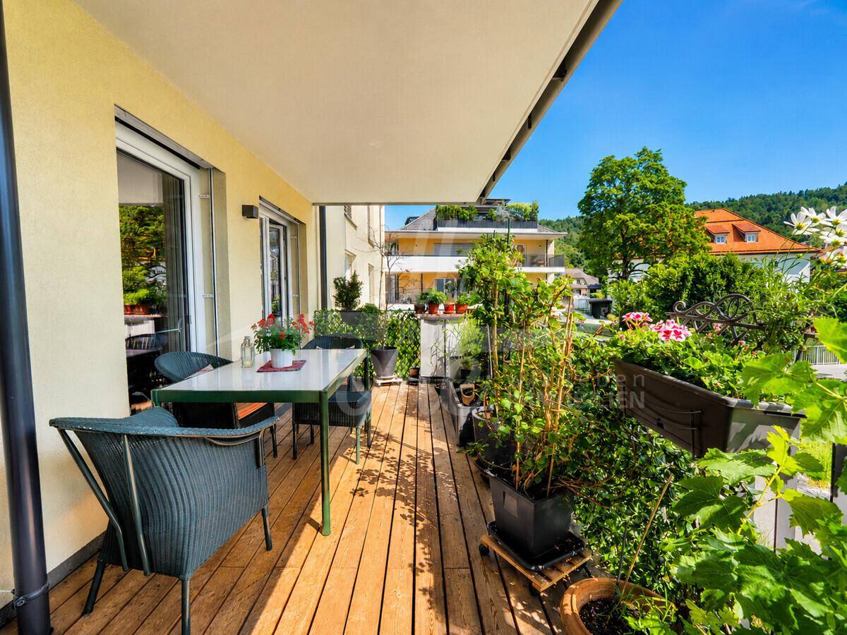 Geräumiger Balkon mit Holzfußboden, vielen Pflanzen und gemütlichen Sitzgelegenheiten im Freien.