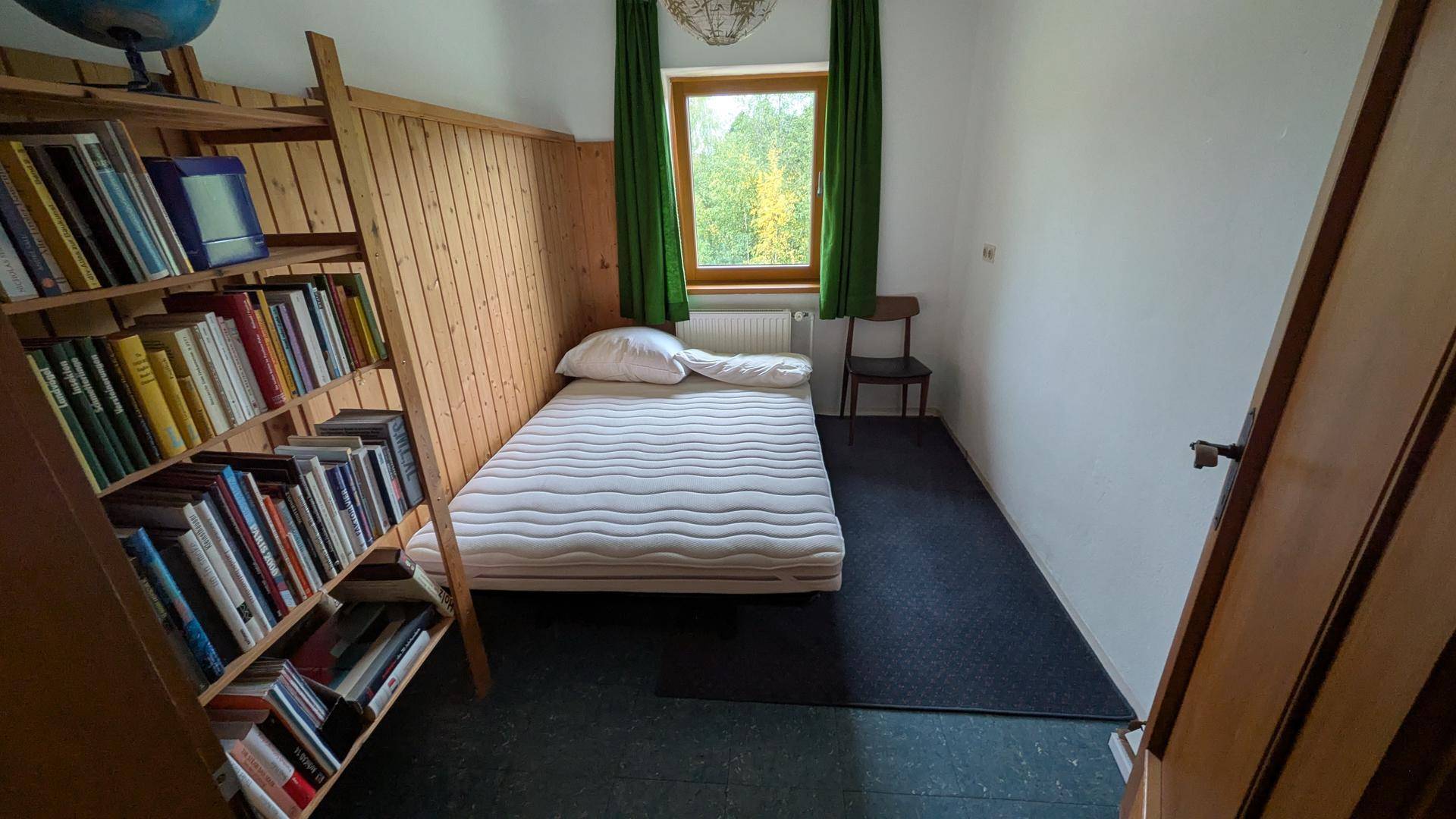Kleines Schlafzimmer mit Einzelbett, Bücherregal und grünen Vorhängen.