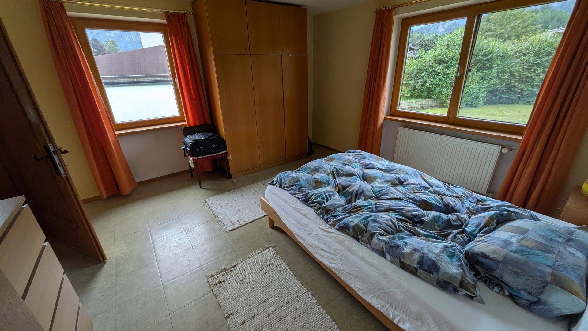 Schlafzimmer mit Einzelbett, Holzschrank und zwei Fenstern mit Ausblick.