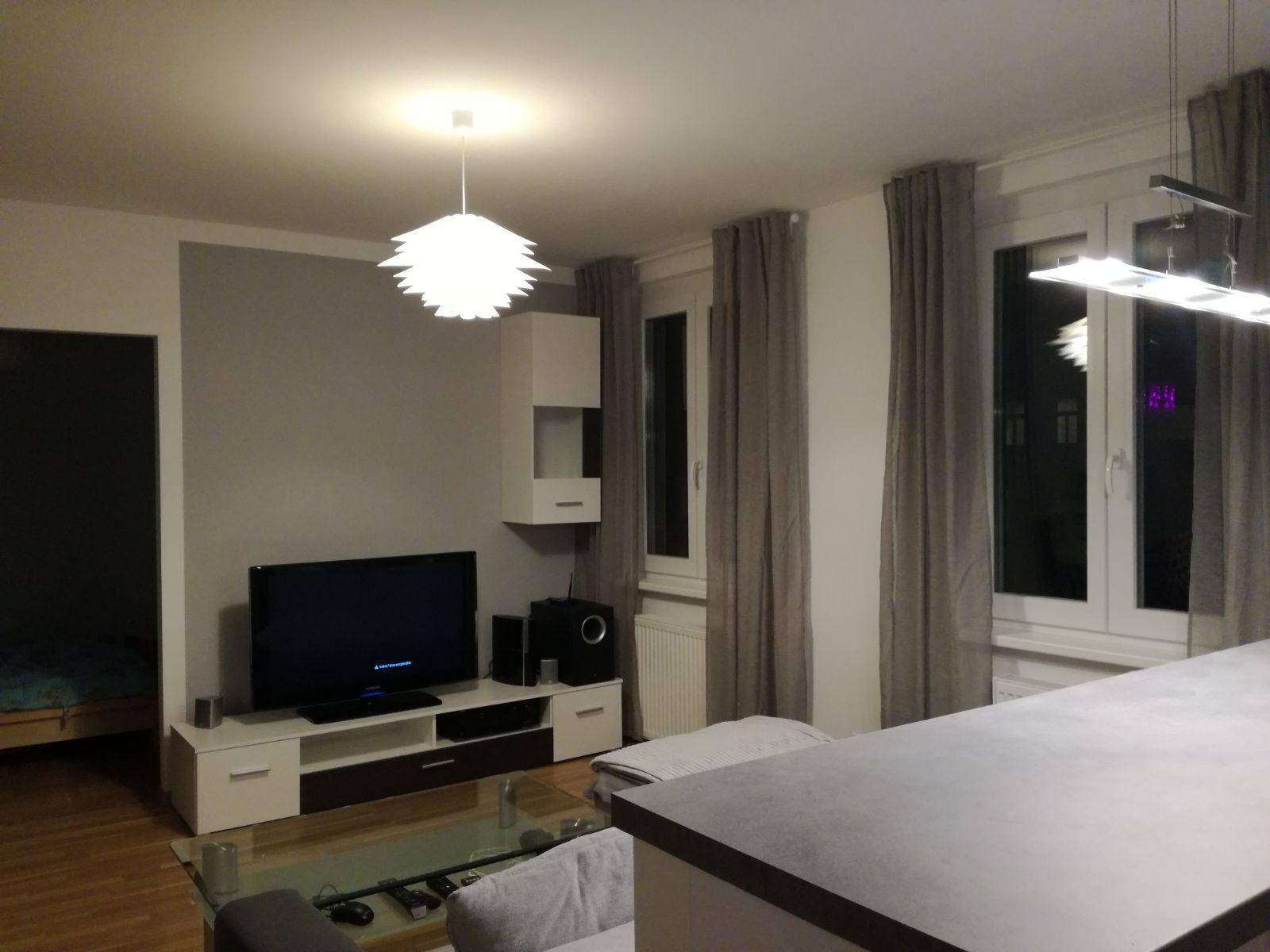 Gemütliches Wohnzimmer mit Fernseher, Sofa und großen Fenstern.