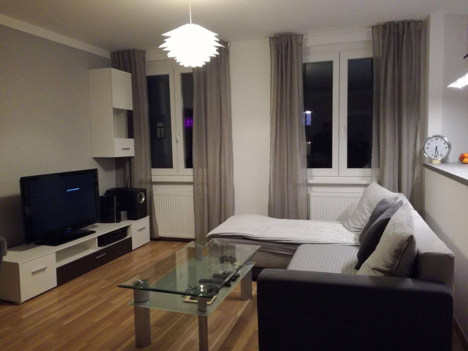 Helles Wohnzimmer mit bequemer Couch, Fernseher und großen Fenstern.