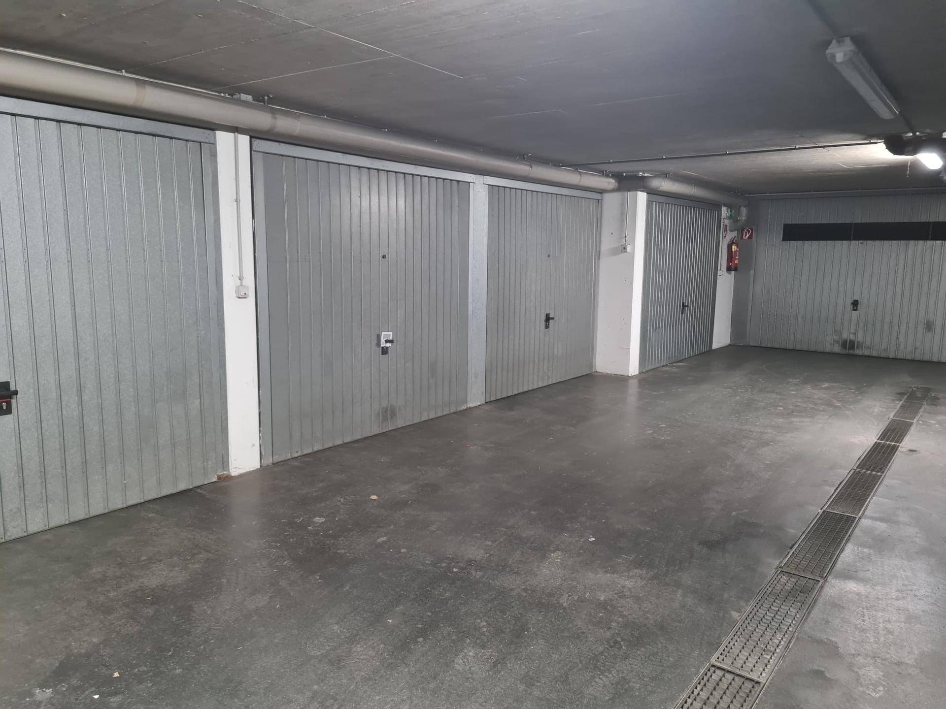 Weitläufige Tiefgarage mit mehreren geschlossenen Garagenboxen und Entwässerungsrinne am Boden.