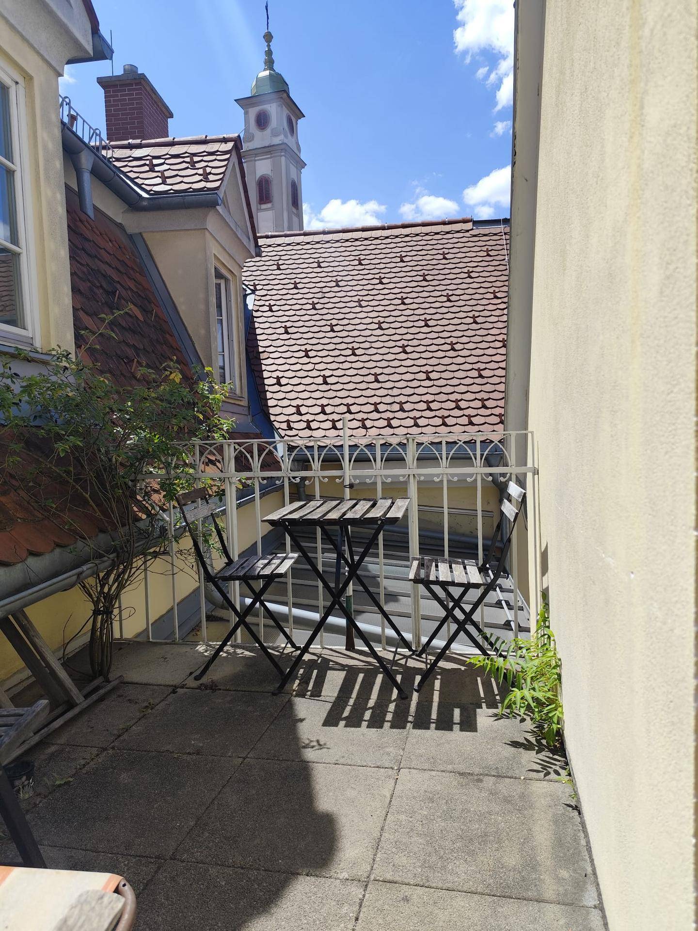 Gemütlicher Balkon mit Tisch und Stühlen, umgeben von traditionellen Dächern und Kirchturm.
