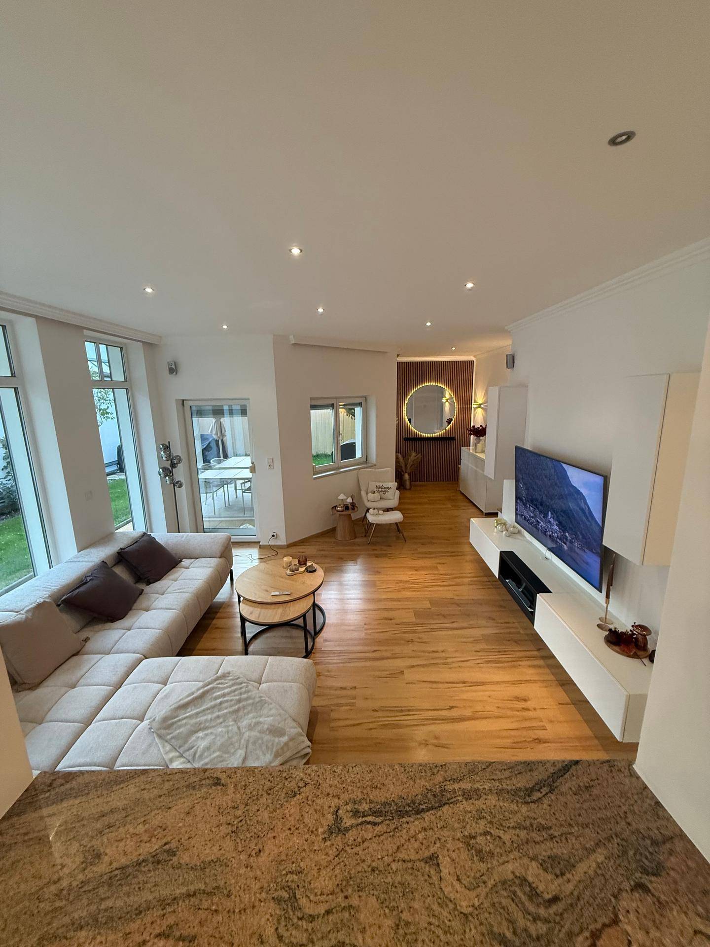 Lichtdurchflutetes Wohnzimmer mit beigem Sofa, Holzboden und Blick ins Grüne.